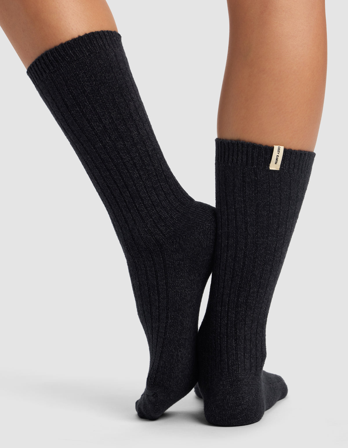 A person stands with feet crossed on a light gray surface, wearing ribbed, dark-colored The Plush Lounge Calf Sock by Cozy Earth with a small fabric tag visible at mid-calf.
