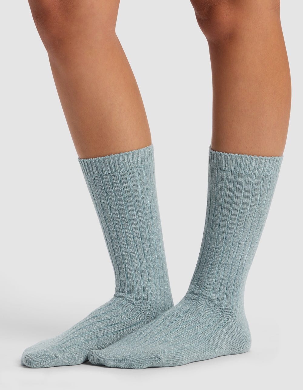 Someone wearing Cozy Earth’s The Plush Lounge Sock 3-Pack in light blue, featuring a ribbed crew design, stands on a white surface, displaying their lower legs and feet.