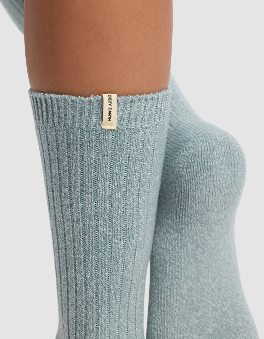 Close-up of lower legs wearing light blue ribbed socks from Cozy Earth's The Plush Lounge Sock 3-Pack, featuring a small beige tag with the brand name. Background is plain white.