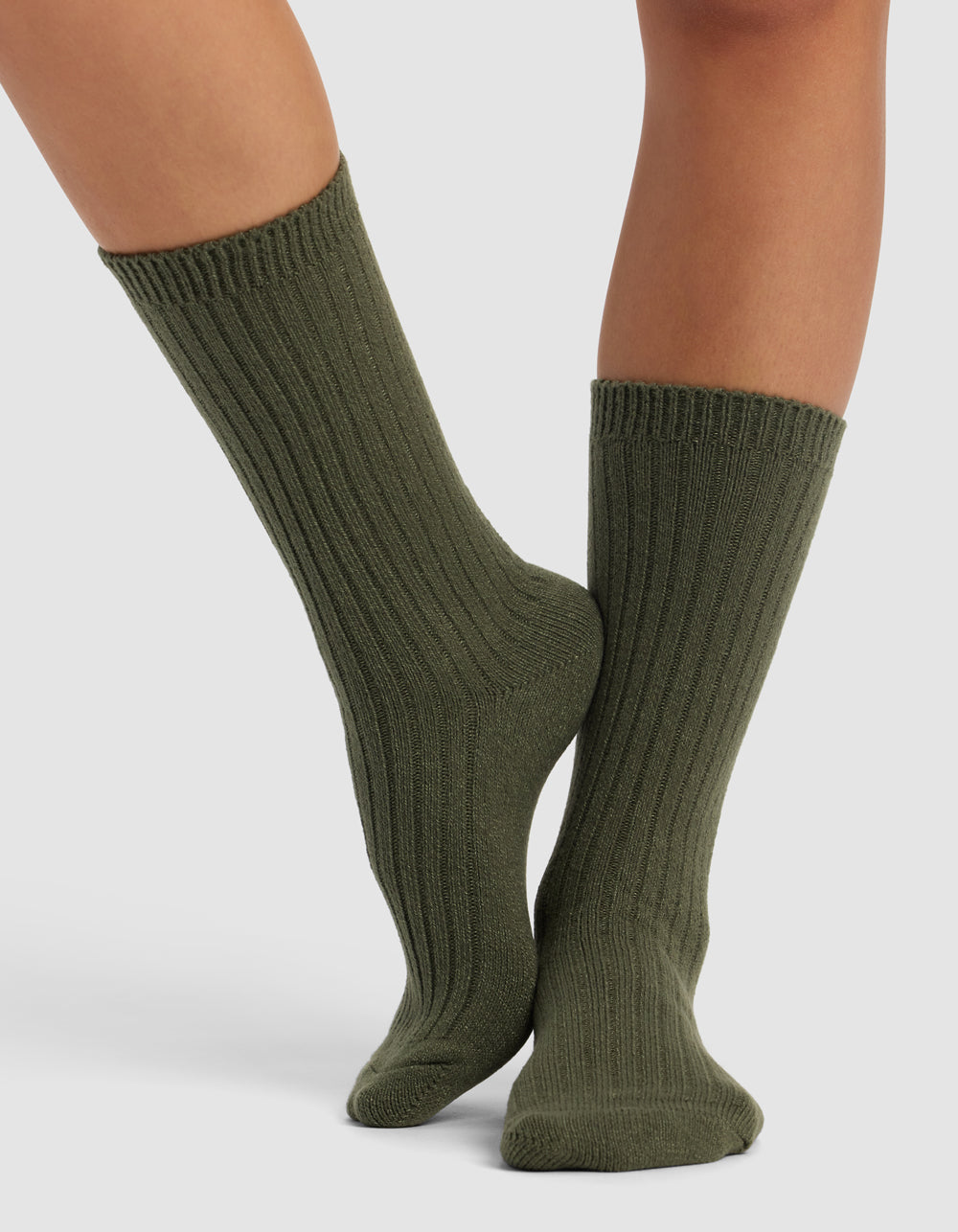 A pair of feet wears Cozy Earth's The Plush Lounge Calf Sock 3-Pack in ribbed olive green, featuring a thick, cozy texture. Legs are crossed at the ankles against a plain, light background.
