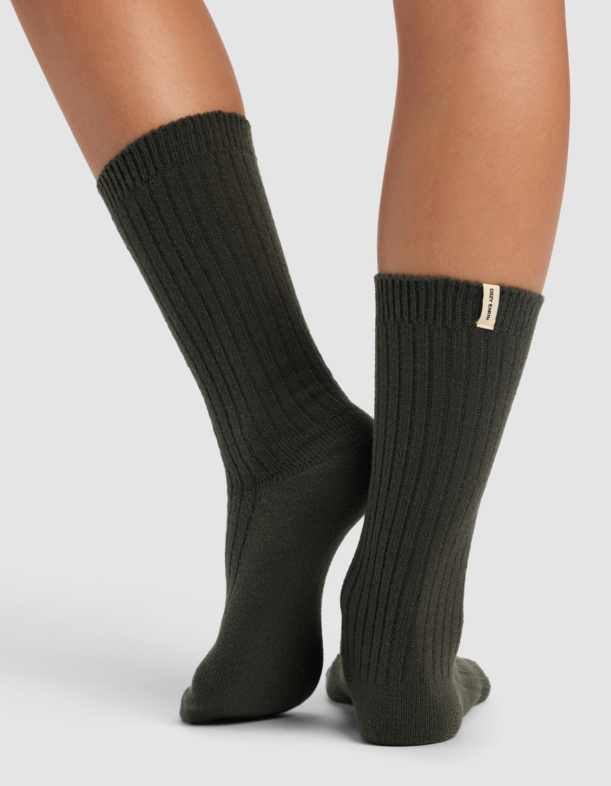 A person stands with crossed legs, wearing Cozy Earth's The Plush Lounge Sock 3-Pack in dark green, featuring ribbed texture and a small beige tag on one cuff. The background is plain light gray.