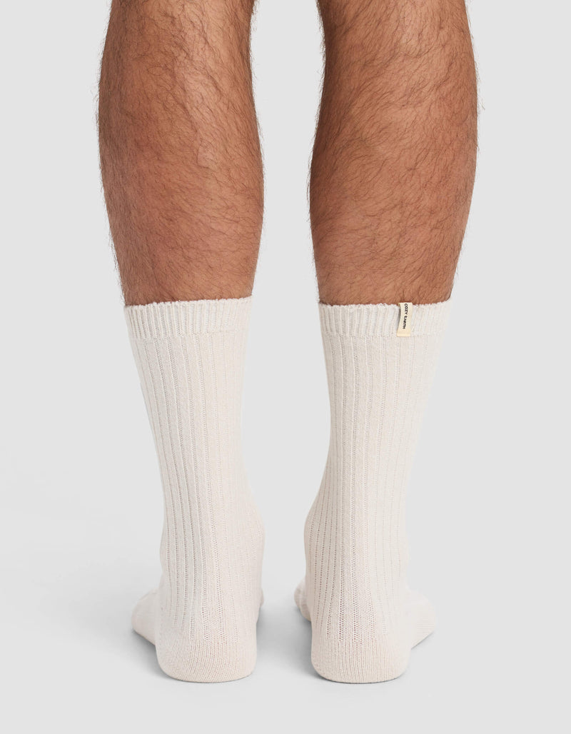 A person’s lower legs are shown from behind, wearing Cozy Earth Men’s Plush Lounge Calf Socks in cream with ribbed texture and a small tag on one sock, set against a plain, light background.