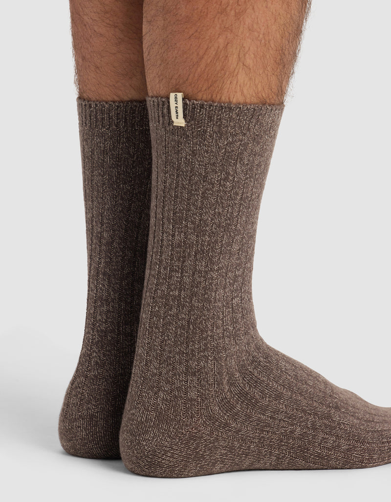 A close-up of two feet wearing thick, ribbed brown socks from Cozy Earth's The Plush Lounge Calf Sock 3-Pack, featuring a small beige tag with text near the top against a plain white background.