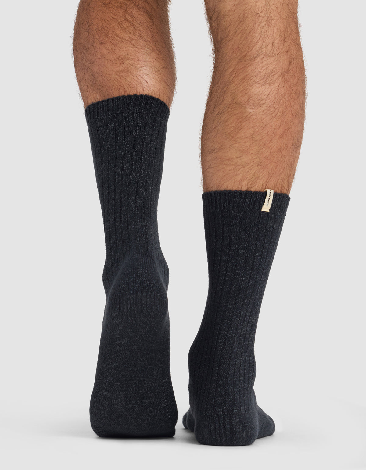 A person in Cozy Earth’s The Plush Lounge Calf Sock 3-Pack stands on a light surface, seen from behind at ankle level. The left heel is slightly raised, and a small tag is visible on the right sock.
