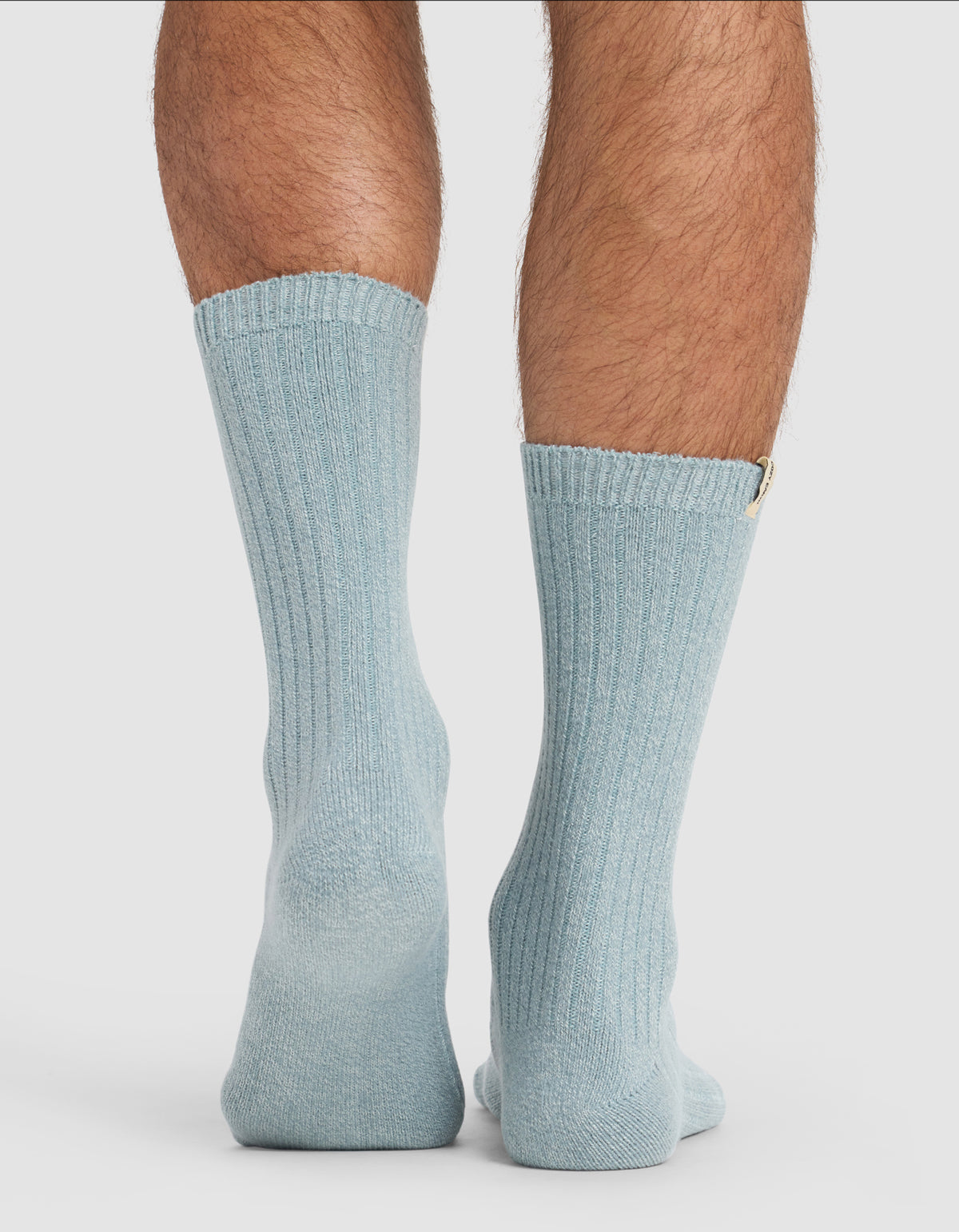 A close-up shows a person’s lower legs wearing Cozy Earth's The Plush Lounge Calf Sock 3-Pack in light blue, standing on a white surface with toes pointed inward.