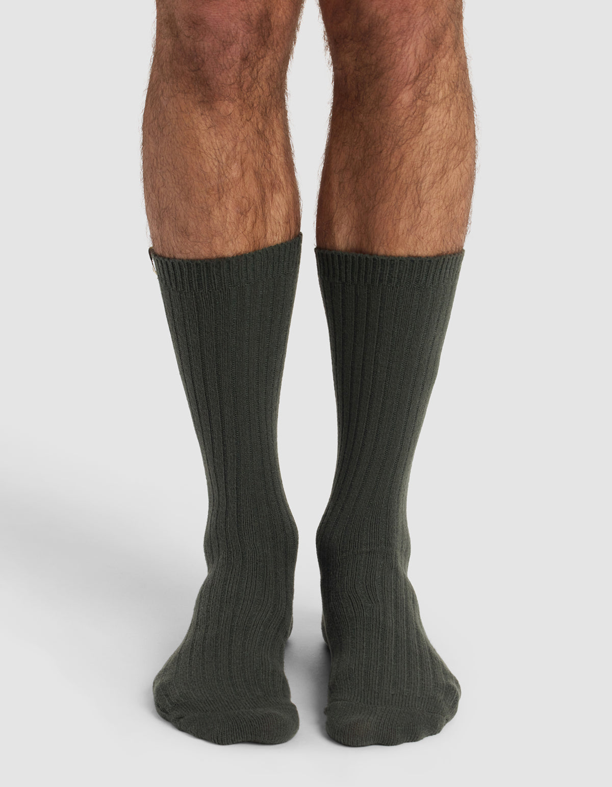 A person with hairy legs stands wearing Cozy Earth’s The Plush Lounge Calf Sock 3-Pack in dark green, shown from the knees down against a plain white background.