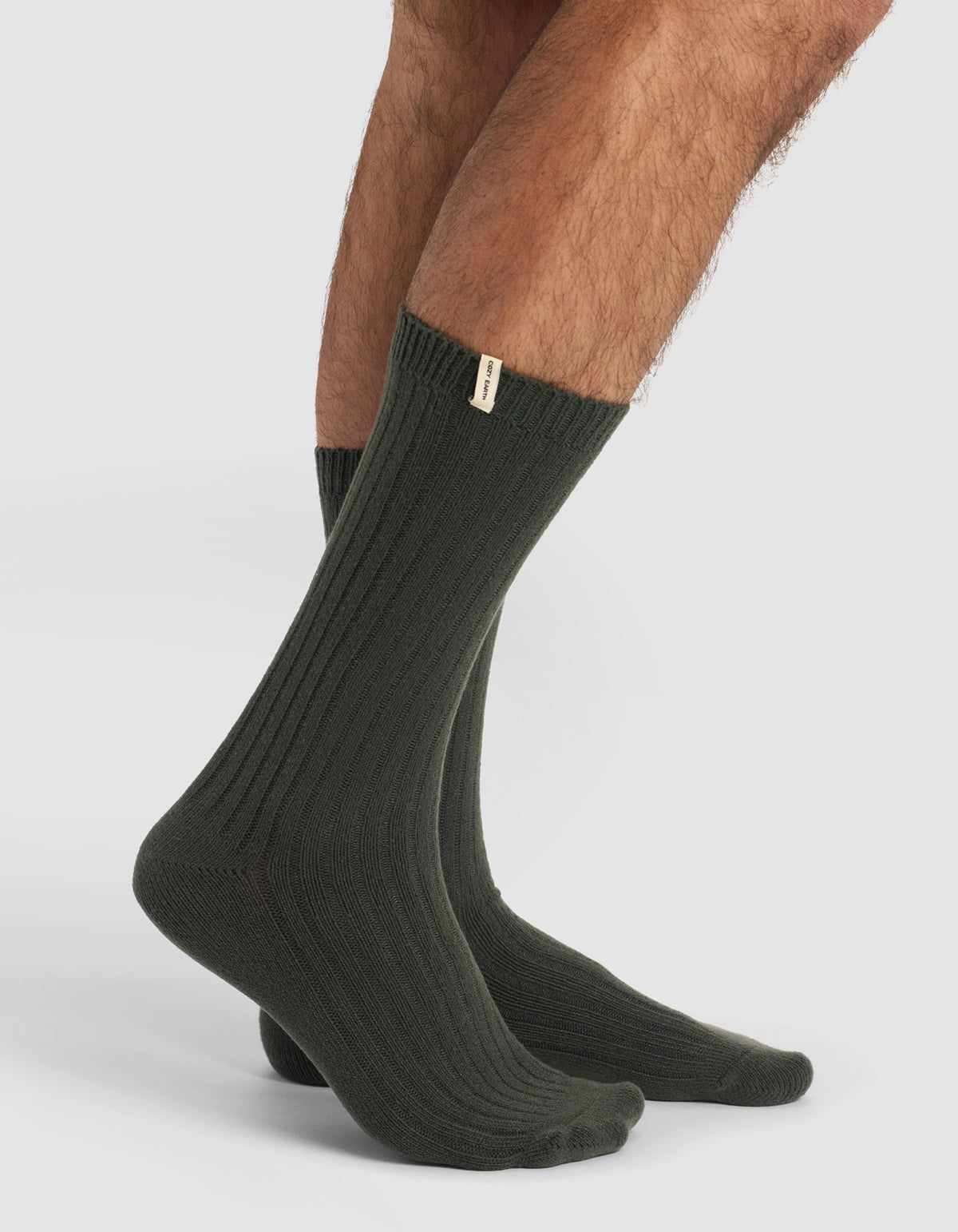 A person wears Cozy Earth’s The Plush Lounge Calf Sock 3-Pack in dark green, featuring ribbed crew style and a small beige tag near the top, standing on a plain white background.