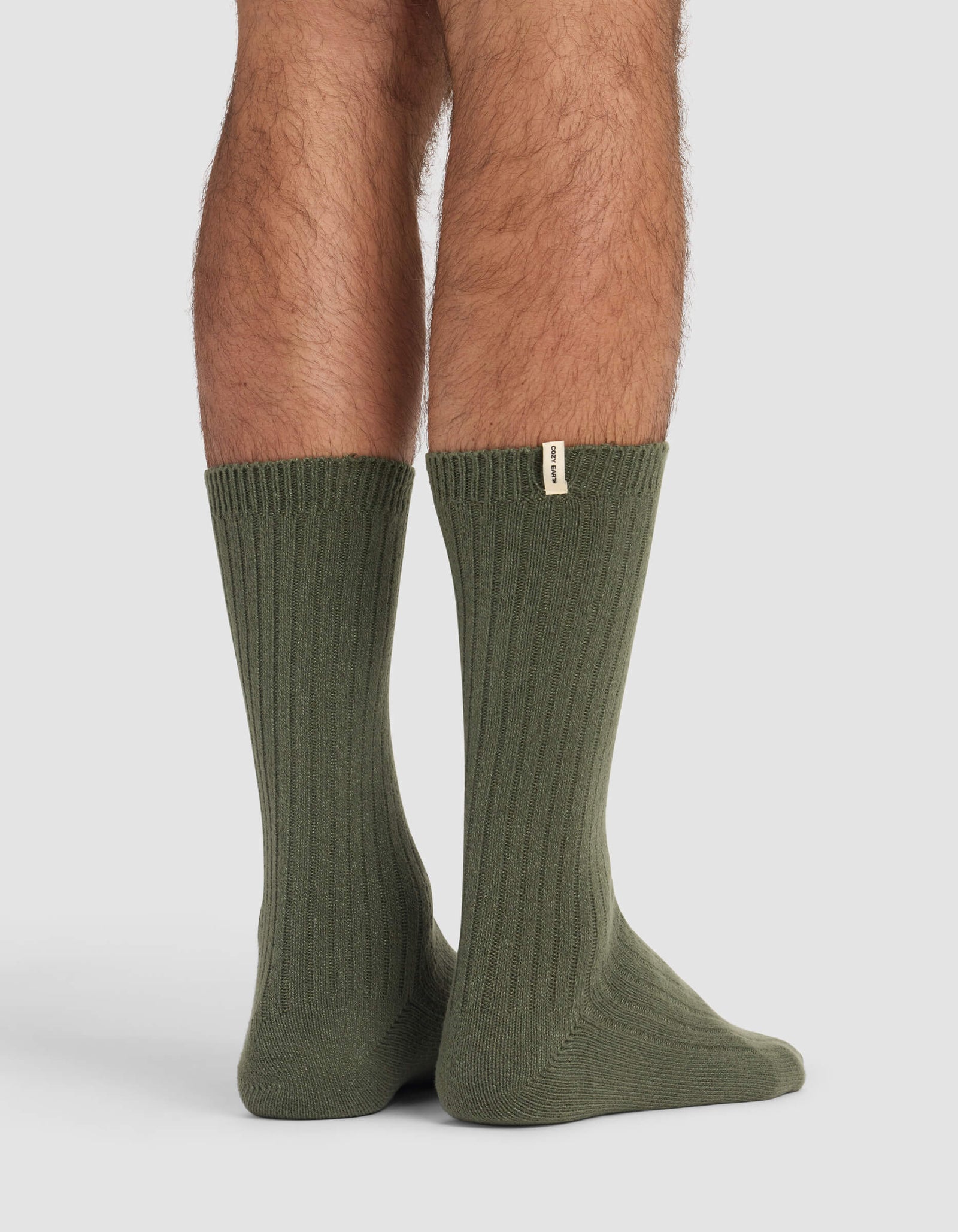 A person stands on a light surface wearing Cozy Earth's Men's Plush Lounge Quarter Sock in olive green, featuring ribbing and a small beige tag near the top. Only their lower legs and feet are visible.