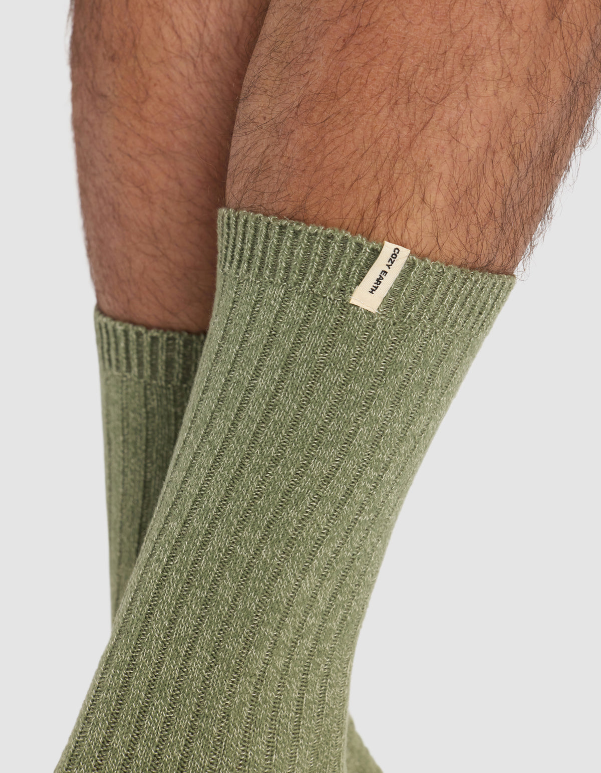 Close-up of a person wearing Cozy Earth's The Plush Lounge Calf Sock 3-Pack in olive green, featuring ribbed texture and a small beige tag near the cuff. Legs appear against a plain, light background.