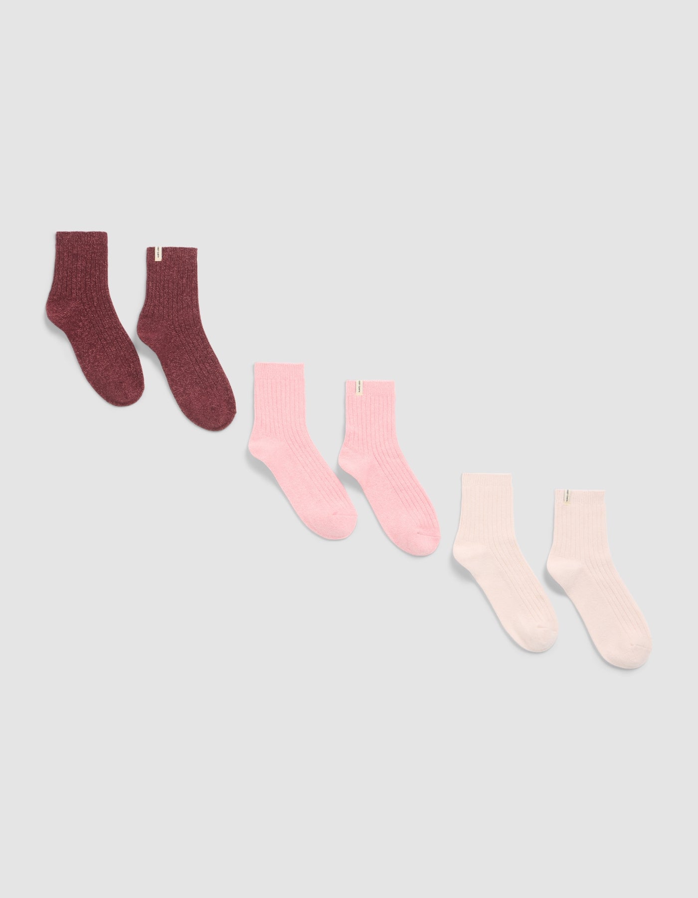 Three pairs of The Plush Modern Crew Sock 3-Pack by Cozy Earth, displayed diagonally on a light background in dark burgundy, pink, and light pink or white shades.