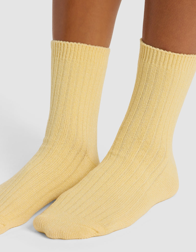 A close-up of lower legs and feet wearing Cozy Earth's The Plush Modern Crew Sock in light yellow, standing on a white surface.