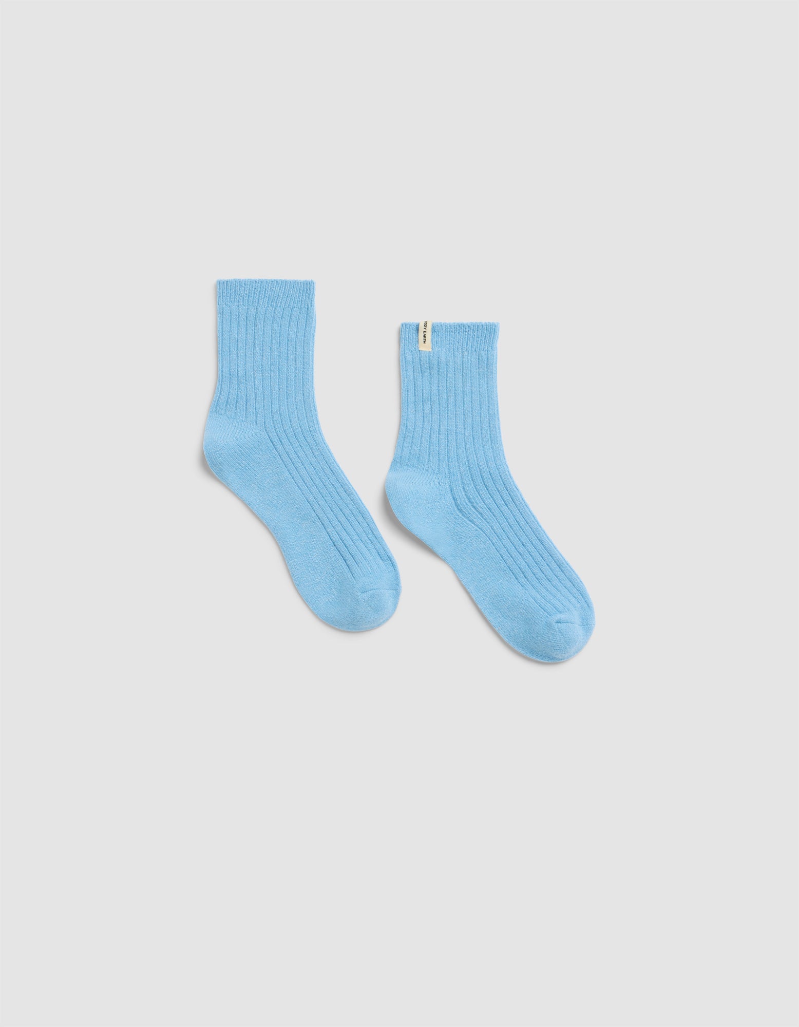The Plush Modern Crew Sock by Cozy Earth in light blue features a ribbed design and a small yellow tag on the upper edge, shown against a plain white background.