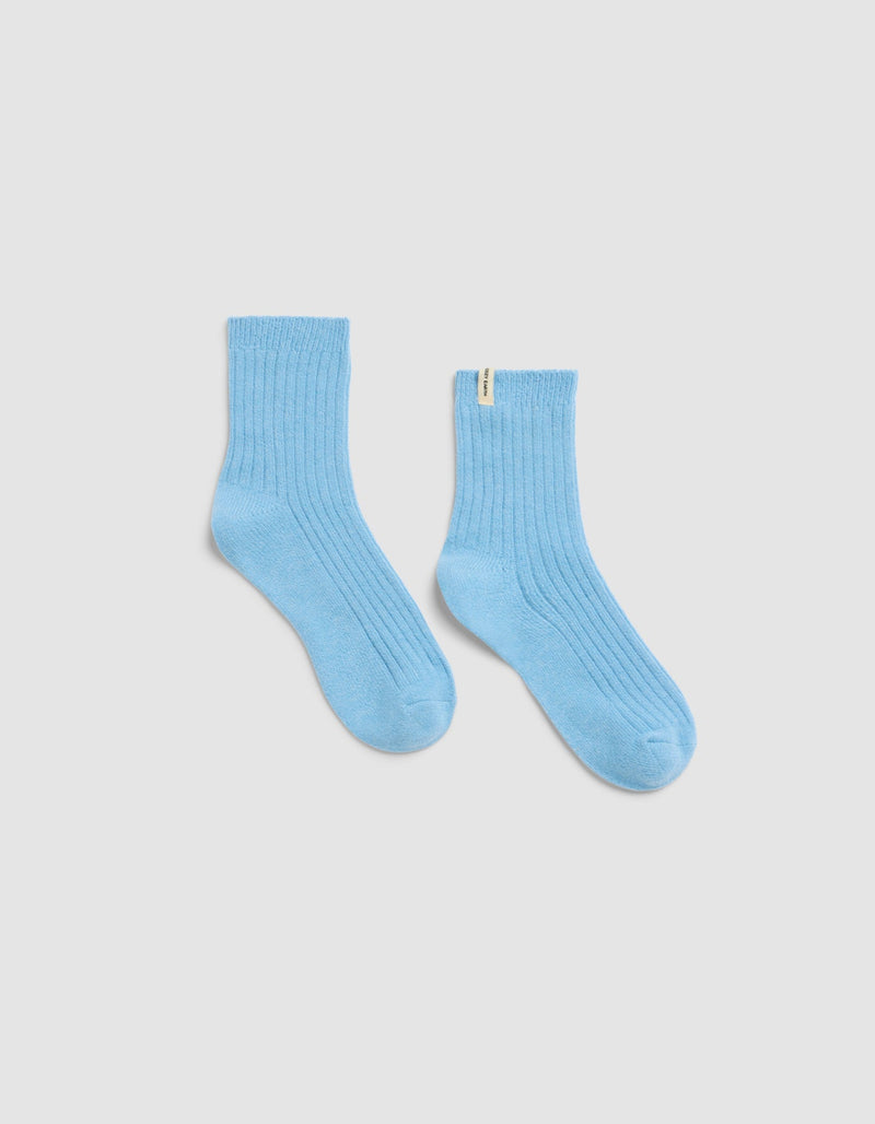 The Plush Lounge Quarter Sock by Cozy Earth in light blue features a ribbed design and a small yellow tag on the upper edge, shown against a plain white background.