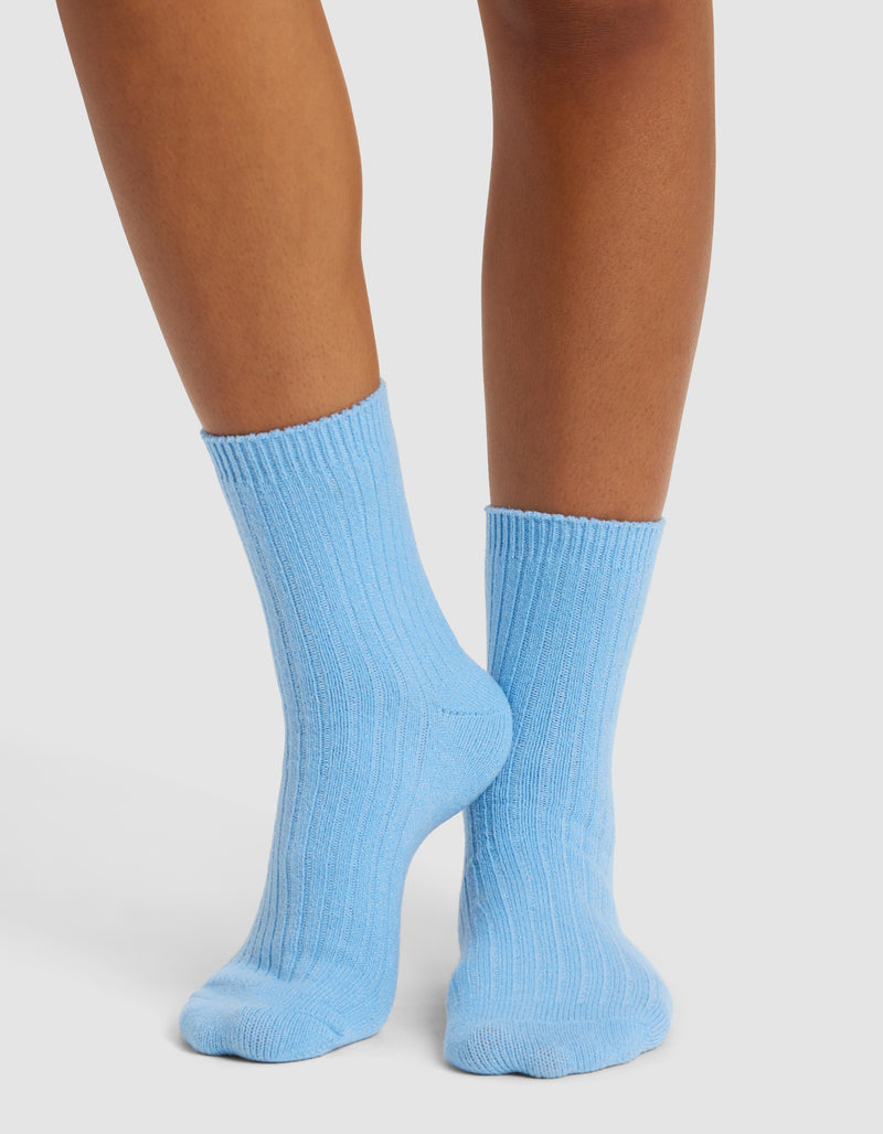 A person wearing Cozy Earth's The Plush Modern Crew Sock in light blue stands on a white surface, with only their lower legs and feet visible.