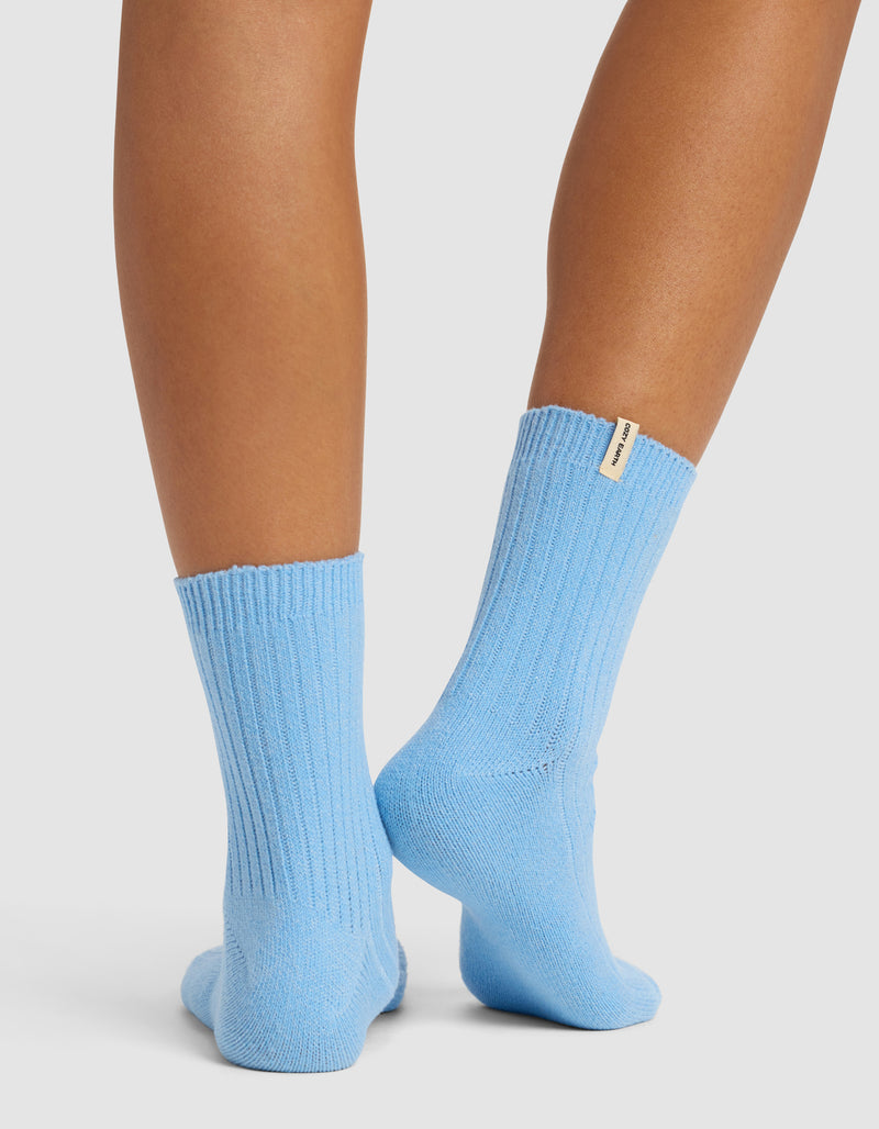 A pair of legs stands on tiptoes, showing the backs of ribbed, light blue Cozy Earth Plush Modern Crew Socks with a small beige tag on one sock against a plain white background.