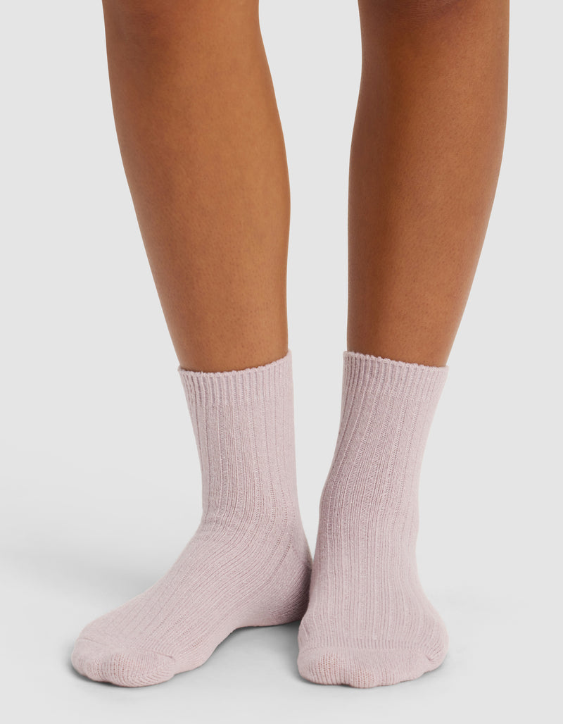 A person wears Cozy Earth’s The Plush Modern Crew Sock in light pink, featuring a ribbed design, standing on a white surface with only their lower legs and feet visible.