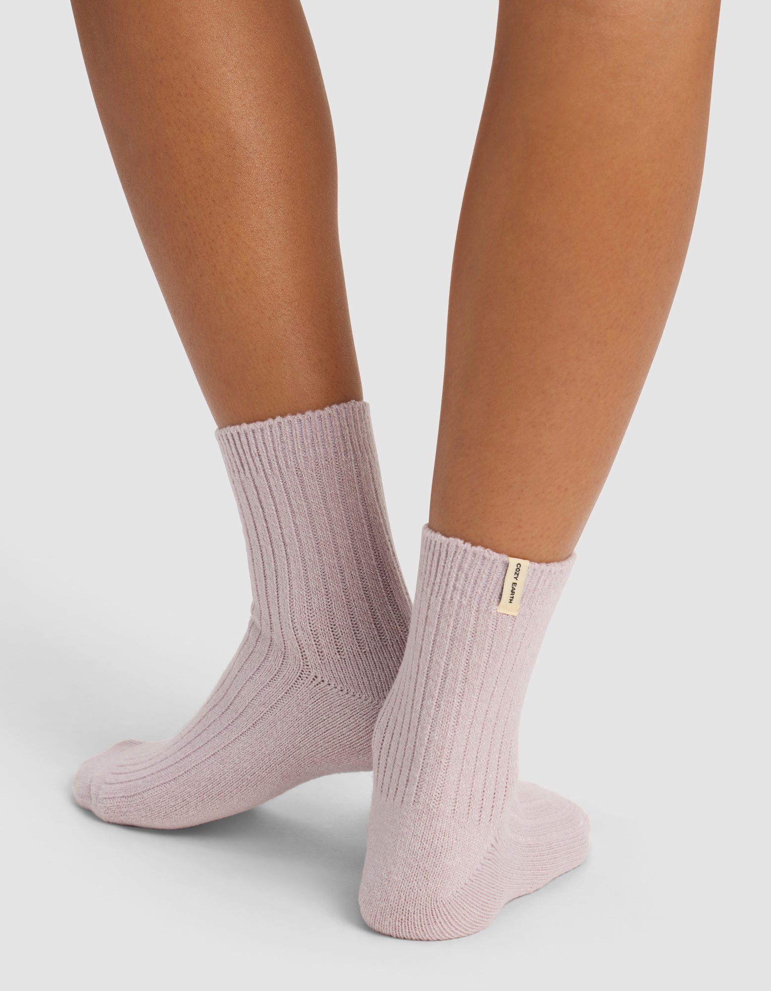 A person stands on tiptoe against a plain background, wearing The Plush Modern Crew Sock by Cozy Earth in light purple ribbed fabric with a small beige tag on one sock.