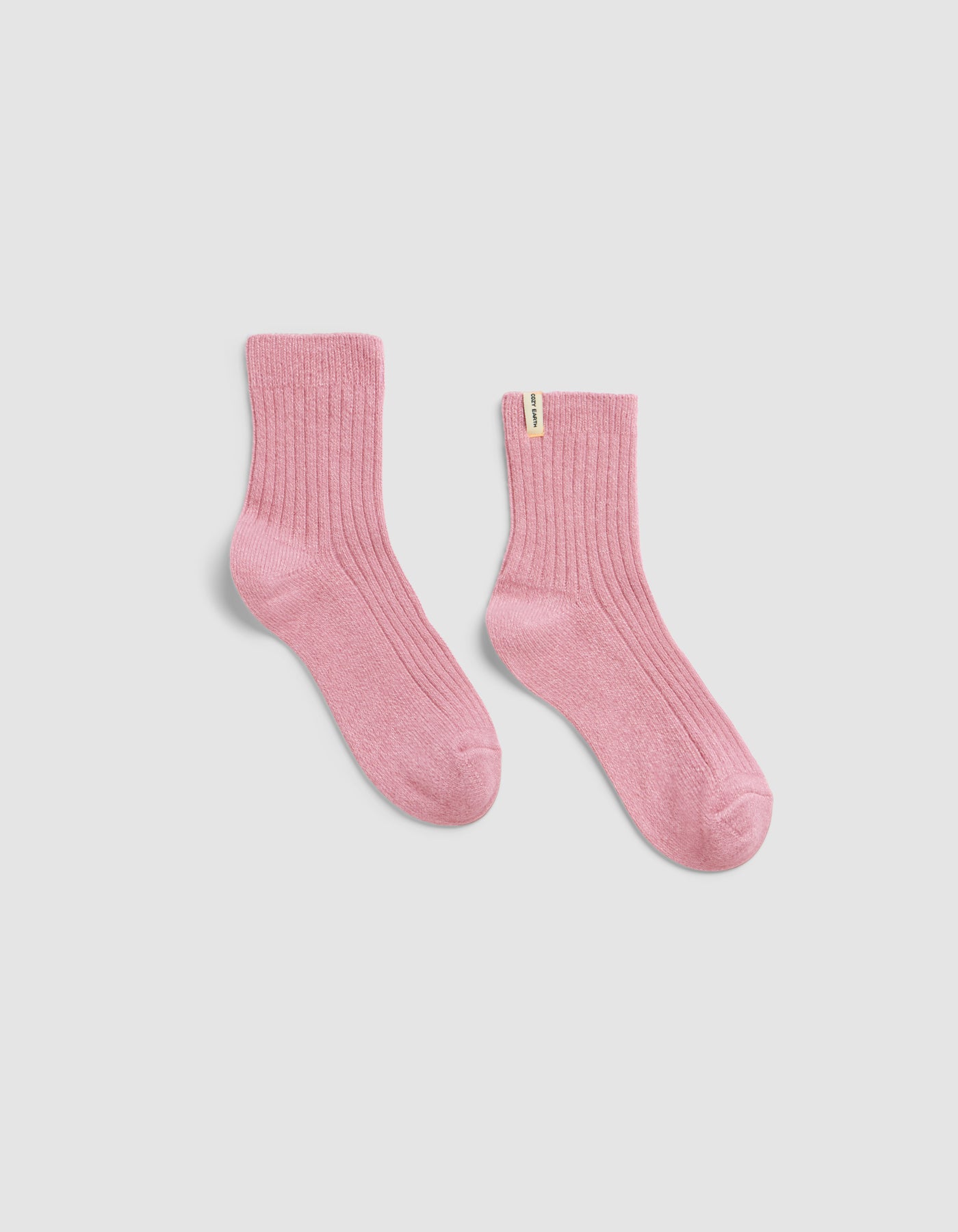 The Plush Modern Crew Sock by Cozy Earth—a pair of ribbed, ankle-length pink socks featuring a small white tag on the top edge—displayed on a light gray background.