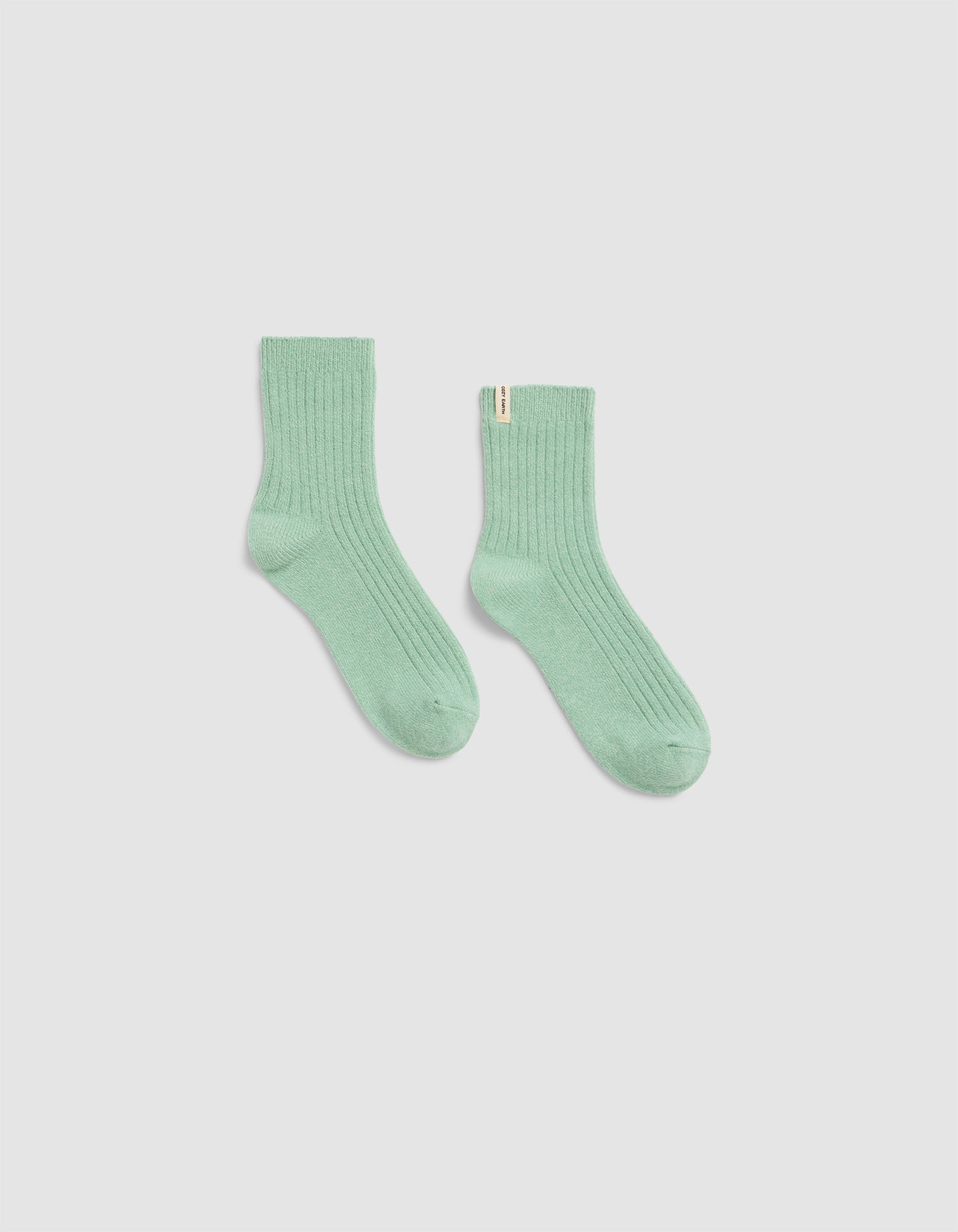 A pair of The Plush Modern Crew Socks by Cozy Earth, in light green ribbed fabric, is laid flat on a plain white background with one sock slightly angled outward.