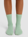 A person with medium brown skin stands on a white surface, wearing Cozy Earth’s The Plush Modern Crew Sock in light green, featuring a ribbed design that covers the ankles and lower calves.|Color:Wintergreen