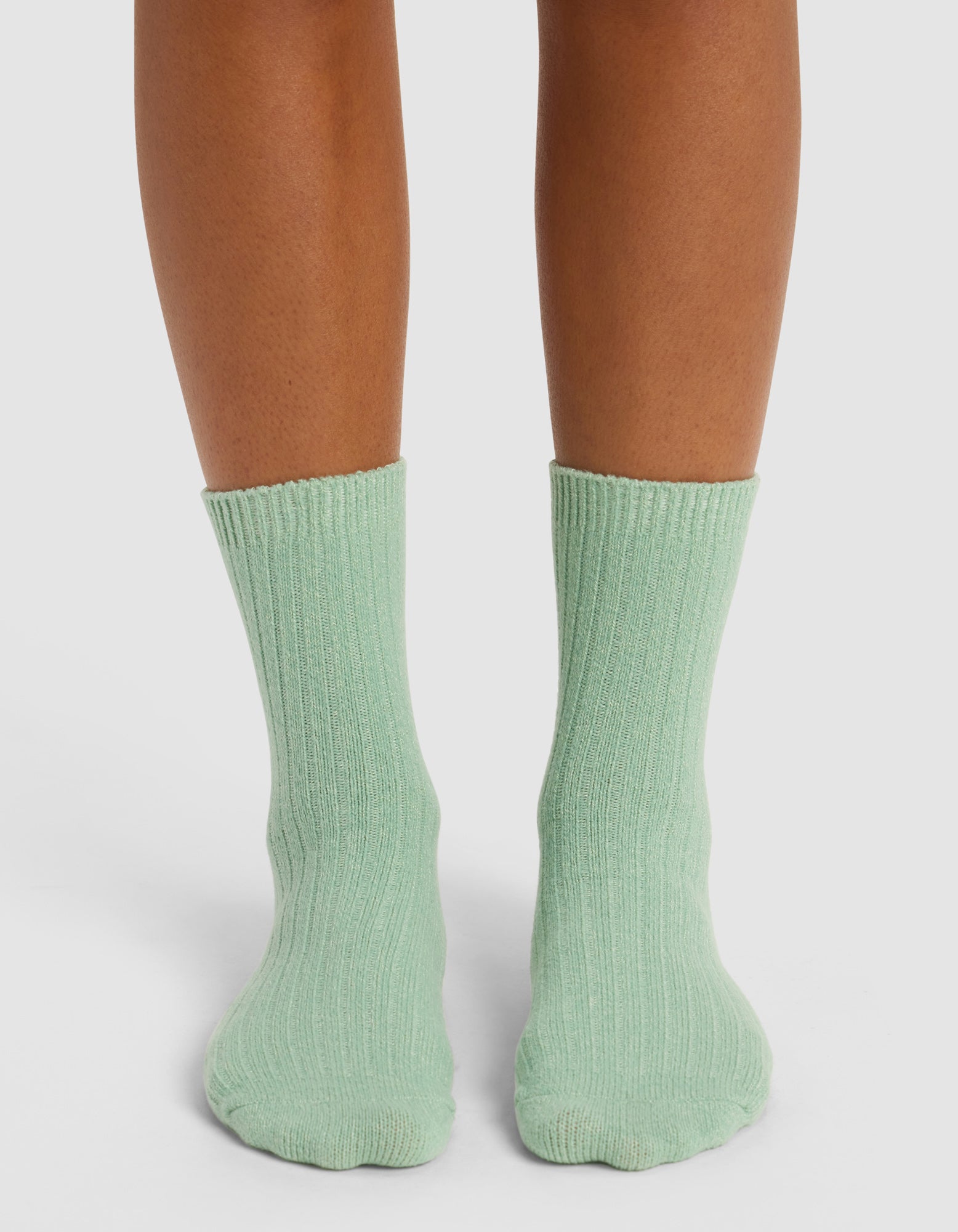 A person with medium brown skin stands on a white surface, wearing Cozy Earth’s The Plush Modern Crew Sock in light green, featuring a ribbed design that covers the ankles and lower calves.|Color:Wintergreen