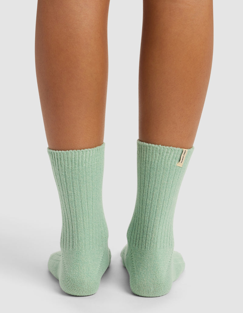 Only the lower legs and feet, seen from behind, show someone standing on a white surface wearing Cozy Earth’s The Plush Modern Crew Sock in mint green ribbed fabric that reaches mid-calf.