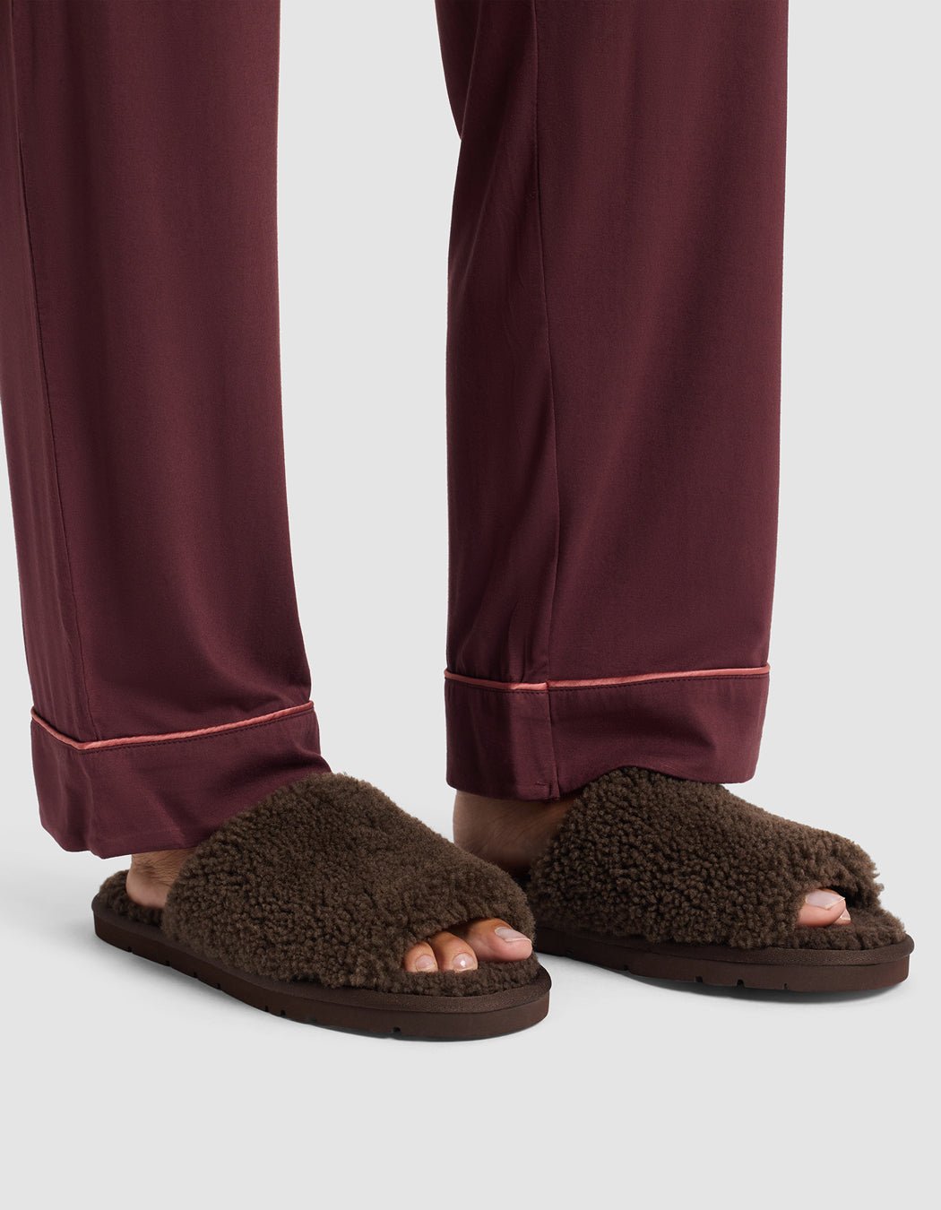 A person wearing burgundy pajama pants with pink piping stands in Cozy Earth Puffy Sheep Slides, brown fuzzy open-toe slippers that show their toes.