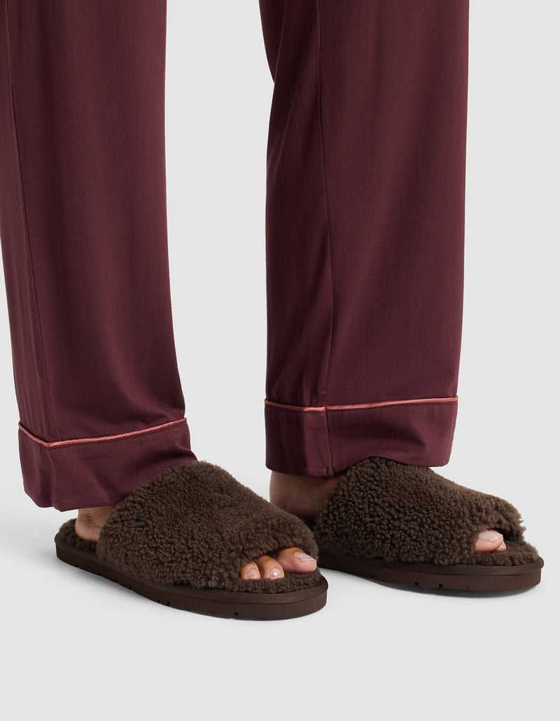 A person wearing burgundy pajama pants with pink piping stands in Cozy Earth Puffy Sheep Slides, brown fuzzy open-toe slippers that show their toes.