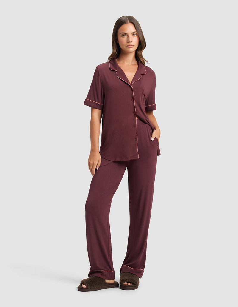 A woman stands facing forward, wearing a maroon pajama set with short sleeves and long pants. She has her left hand in her pocket and is wearing Cozy Earth Puffy Sheep Slides. The background is plain light gray.