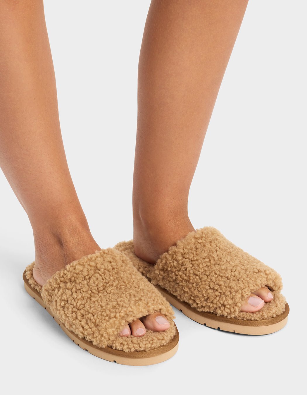 A person wears Cozy Earth’s Puffy Sheep Slides—fuzzy, tan open-toe slippers with thick, textured uppers and flat soles—standing on a white background.
