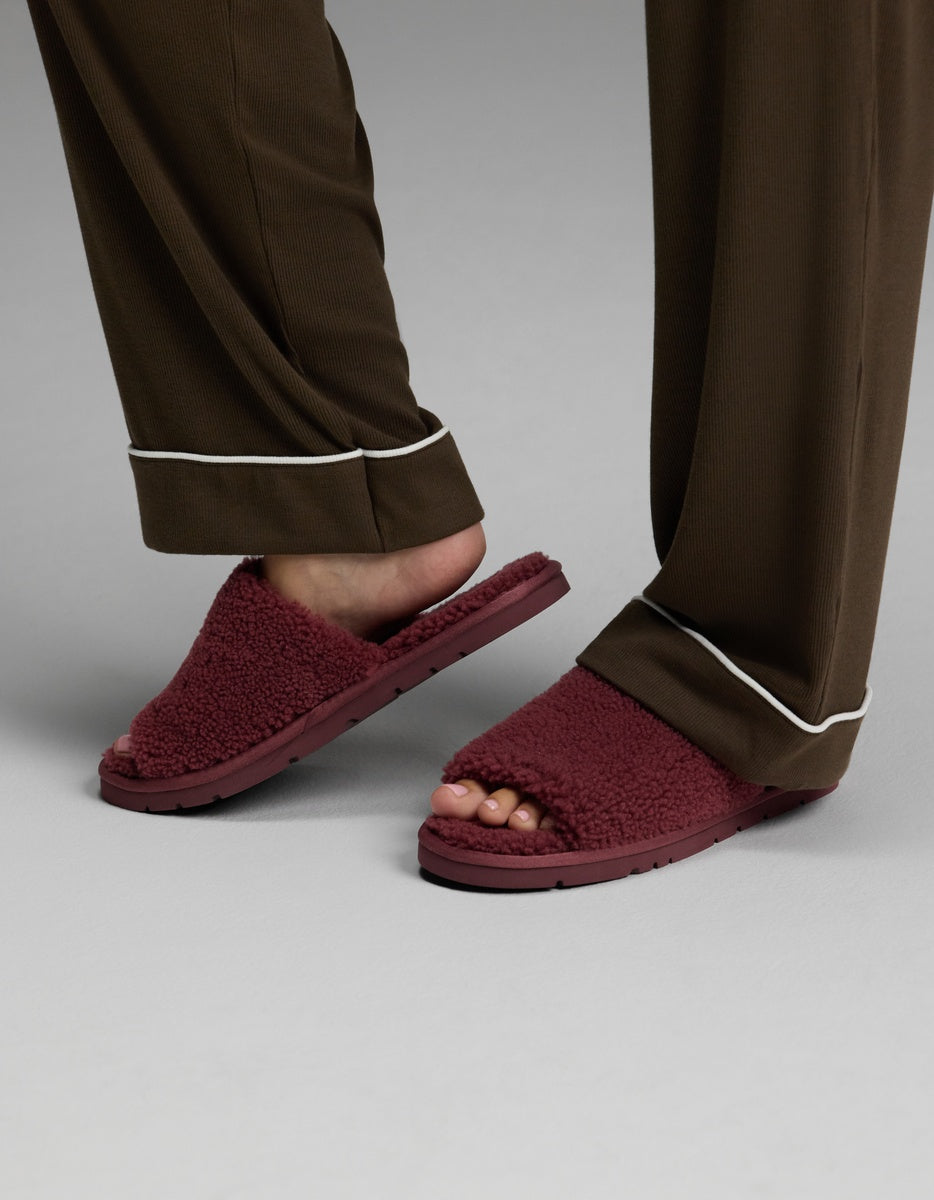 A person is wearing Cozy Earth’s brown pajama pants with white trim and Puffy Sheep Slides—maroon fuzzy open-toe slippers—while standing on a gray surface.
