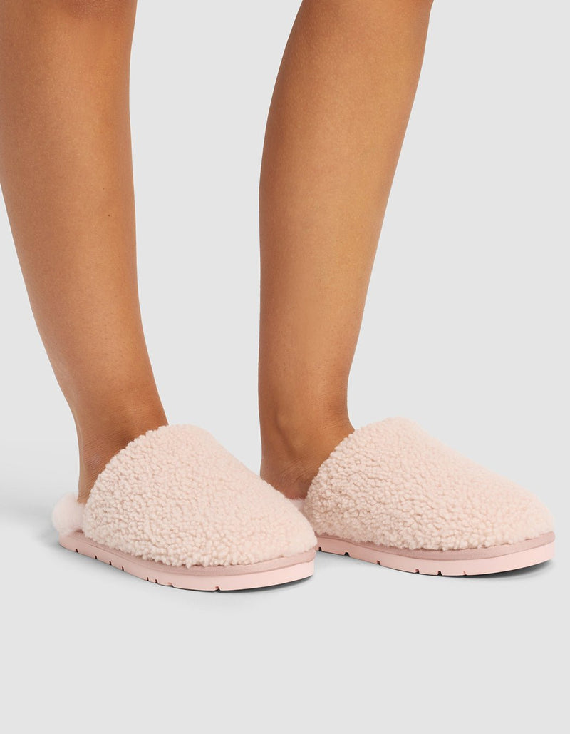 A person wears Cozy Earth Puffy Sheep Slippers in light pink, featuring a soft, sherpa-like texture and flat sole, while standing on a light gray surface. Only their lower legs and feet are visible.