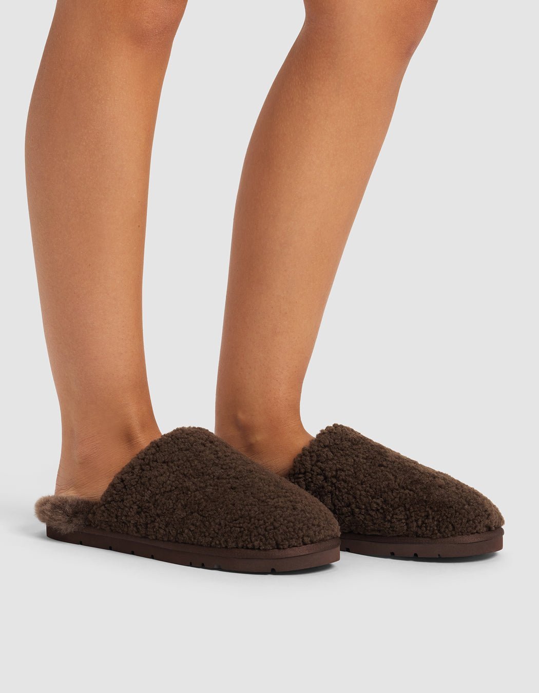 A person wears Cozy Earth Puffy Sheep Slippers—dark brown, fuzzy, slip-on flats. Only their lower legs and feet are visible against a light gray background.