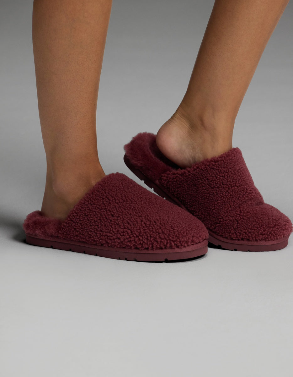 A person wearing Cozy Earth Puffy Sheep Slippers in dark red stands on a light gray surface. The slippers feature a plush, textured design with open heels.