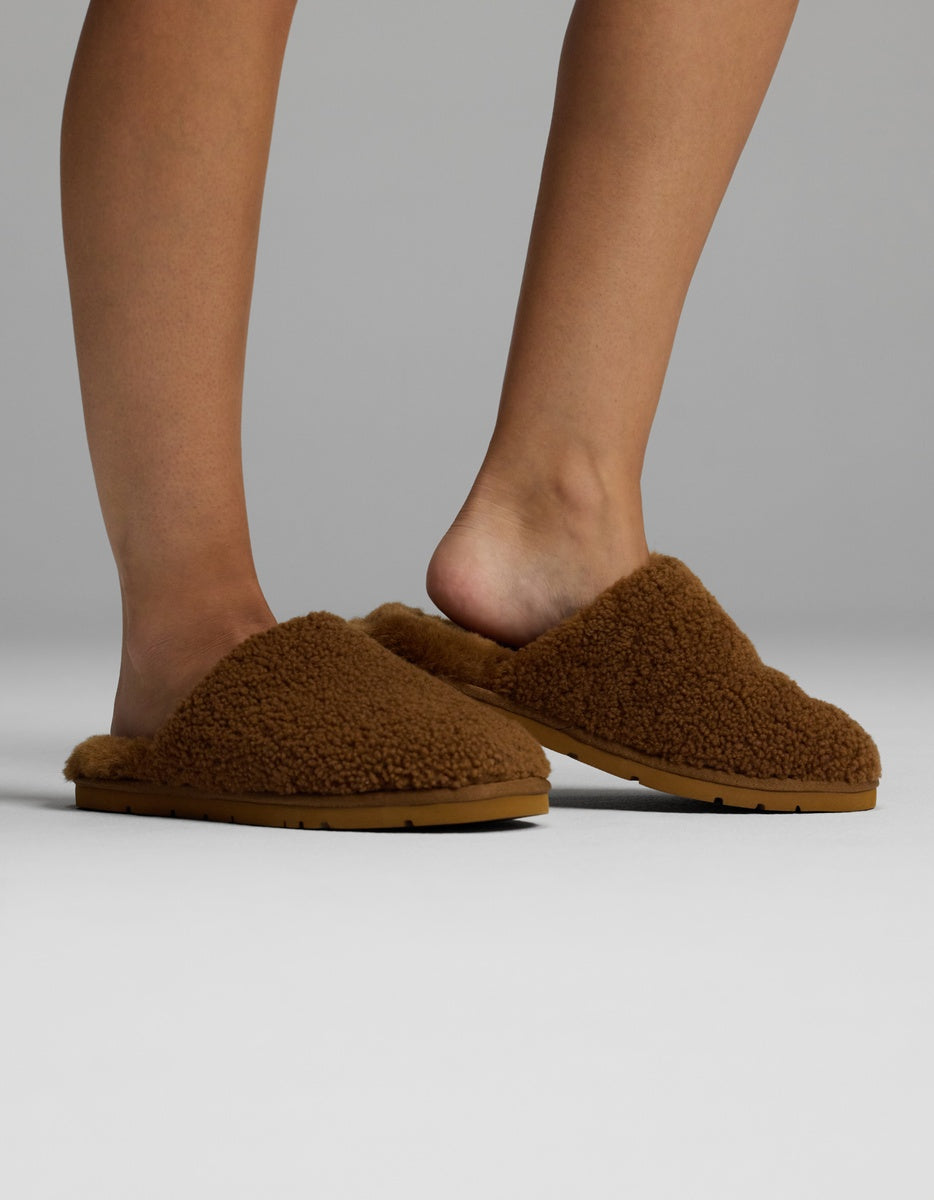 A person wearing Cozy Earth Puffy Sheep Slippers stands on a light grey surface, with one foot slightly raised to reveal the back of the slipper and the other resting flat on the ground.