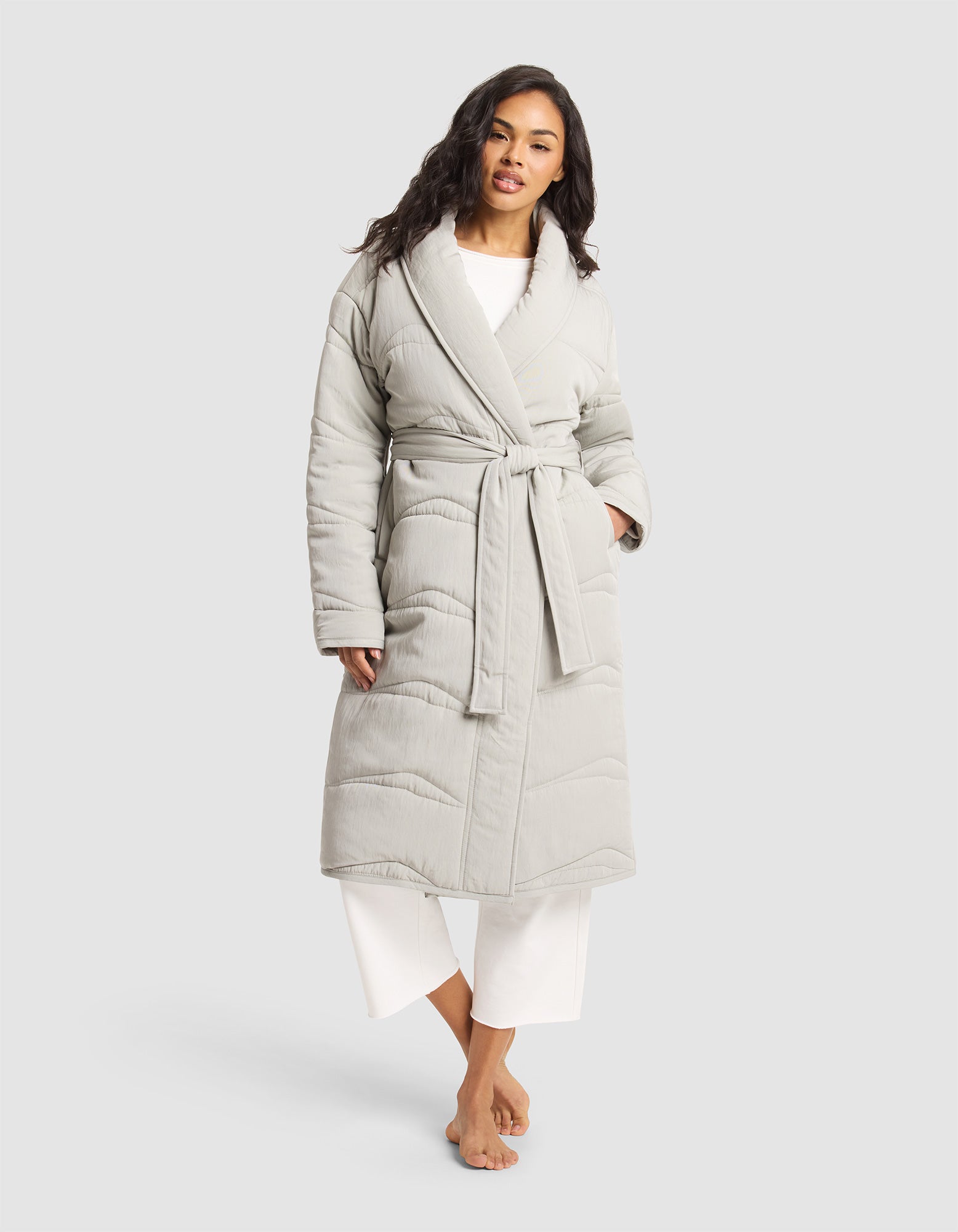 A woman stands barefoot against a plain light background, wearing Cozy Earth's Women's Quilted House Coat in light gray with a tied belt, layered over white pajama pants. |Color:Aloe