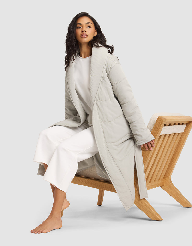 A woman with long dark hair sits barefoot on a wooden chair, wearing the Cozy Earth Women's Quilted House Coat over a white top and wide-legged white pants, against a plain light background.