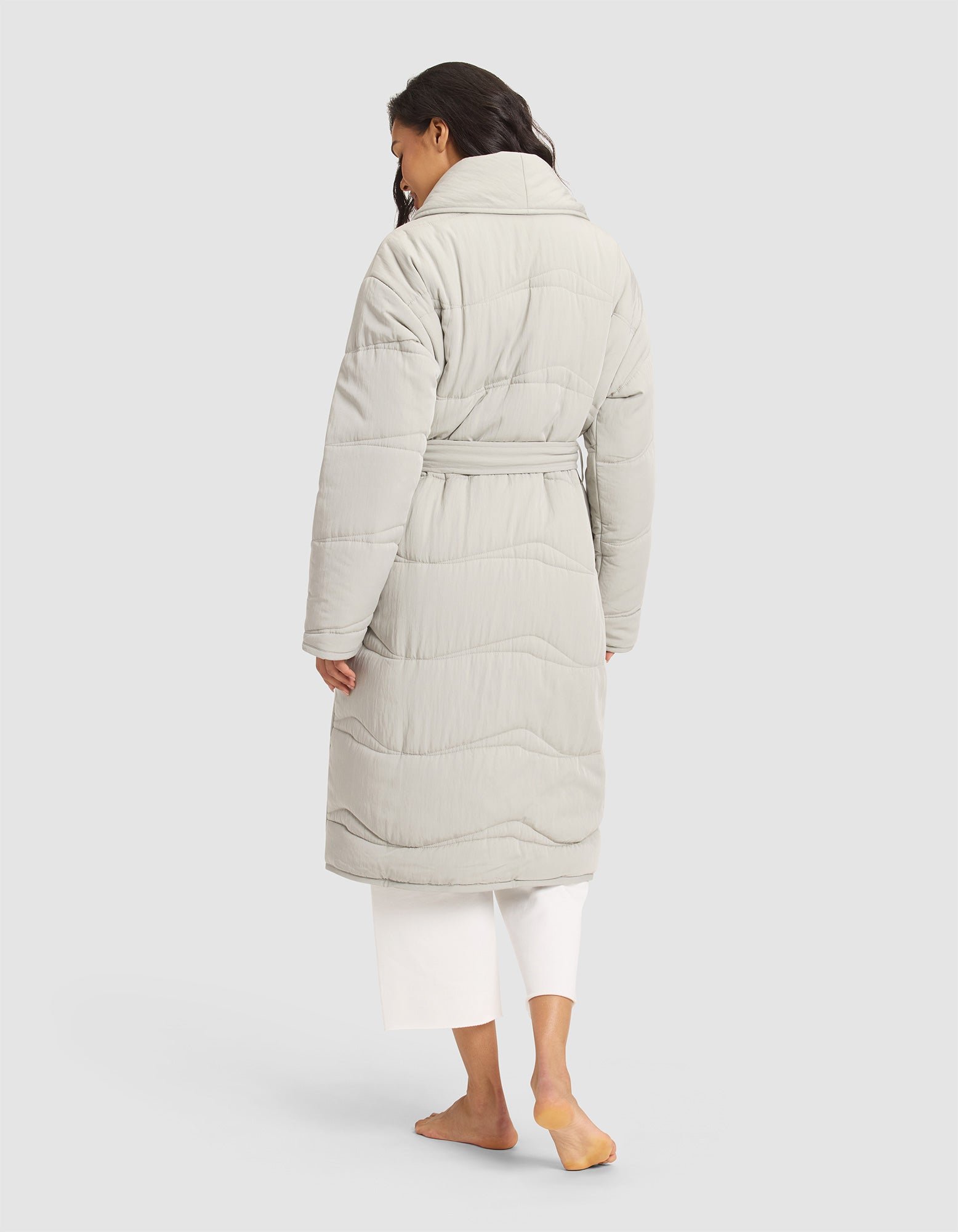 A woman with long, wavy dark hair models the Cozy Earth Women's Quilted House Coat in light gray, paired with white pants. She stands barefoot with her back to the camera against a plain background. 