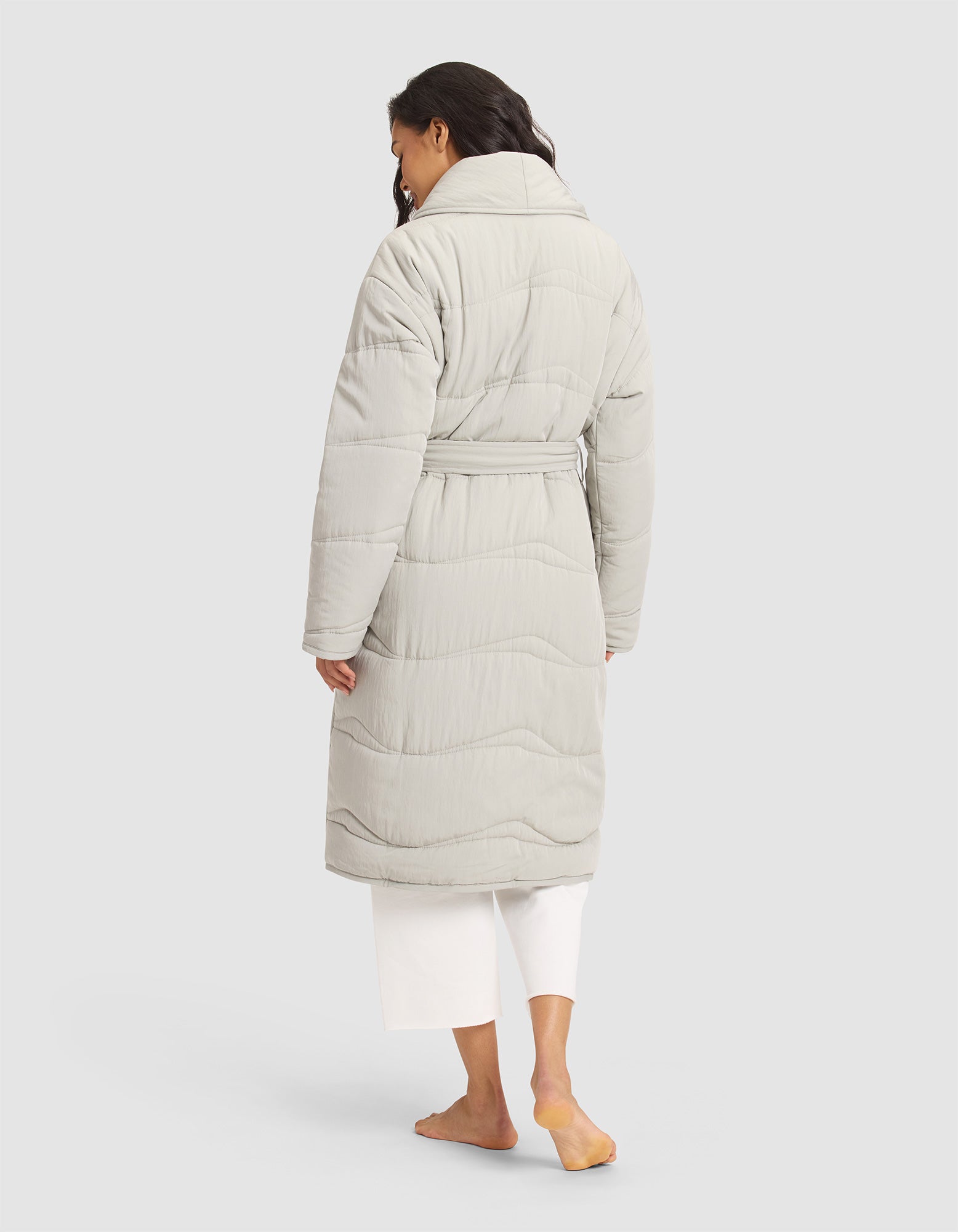 A woman with long, wavy dark hair models the Cozy Earth Women's Quilted House Coat in light gray, paired with white pants. She stands barefoot with her back to the camera against a plain background. 