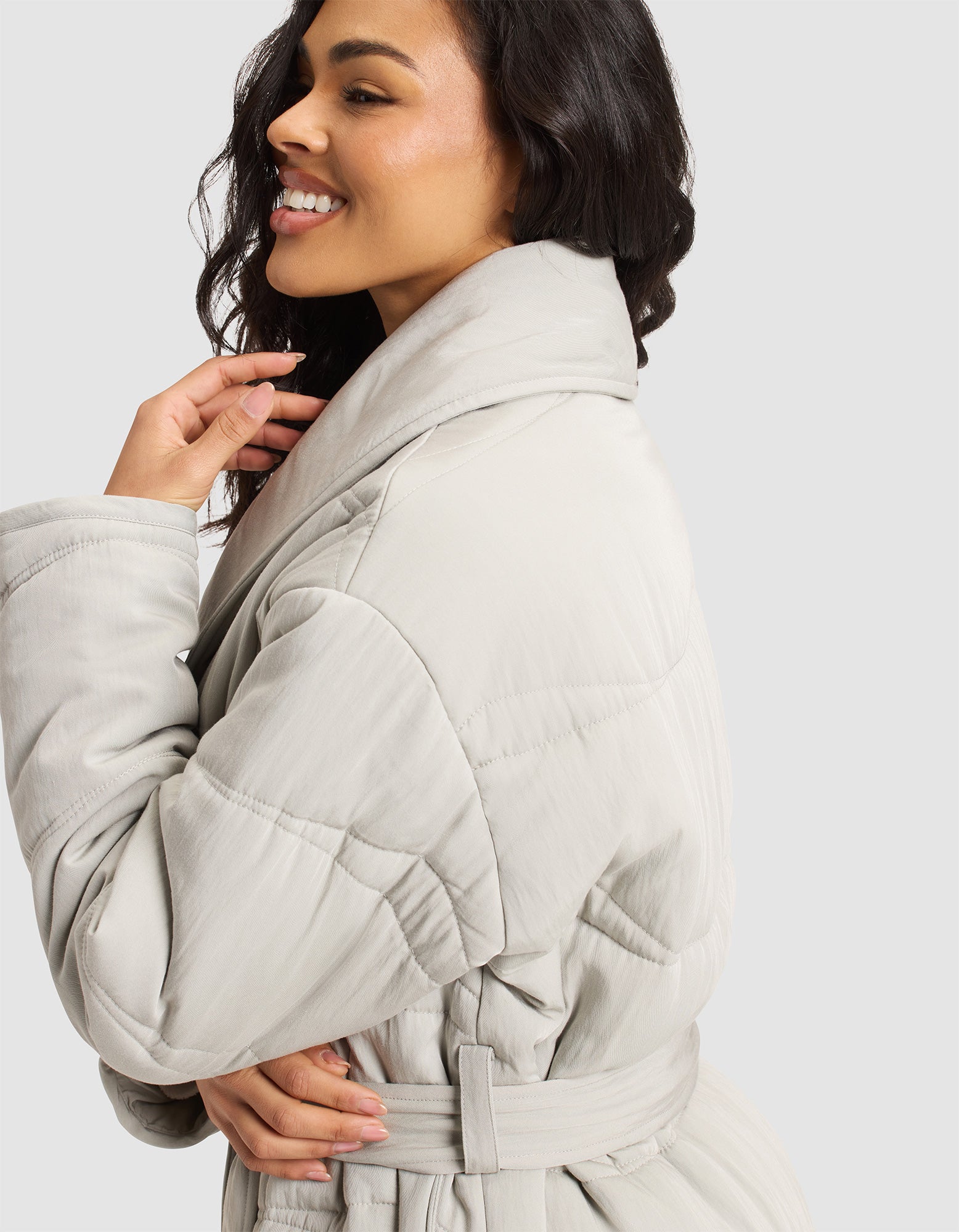 A woman with wavy dark hair smiles in a Cozy Earth Women's Quilted House Coat, light gray with a wide collar and belt, shown from the side against a plain light background. 