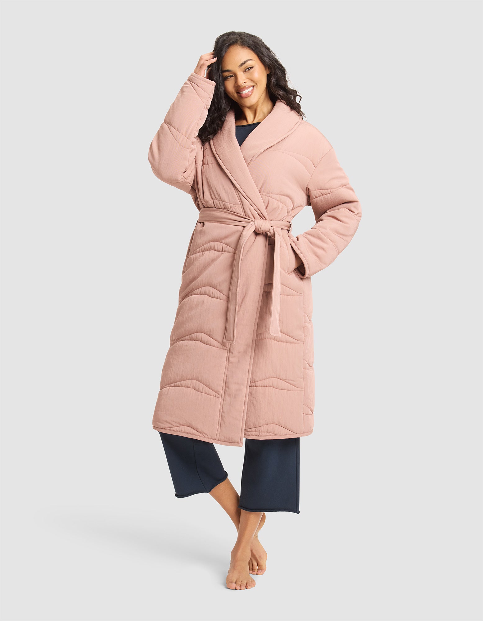 A smiling woman poses barefoot in Cozy Earth’s Women’s Quilted House Coat in Rosewood, belted over dark blue pajama pants, with one hand touching her hair against a light gray background. 