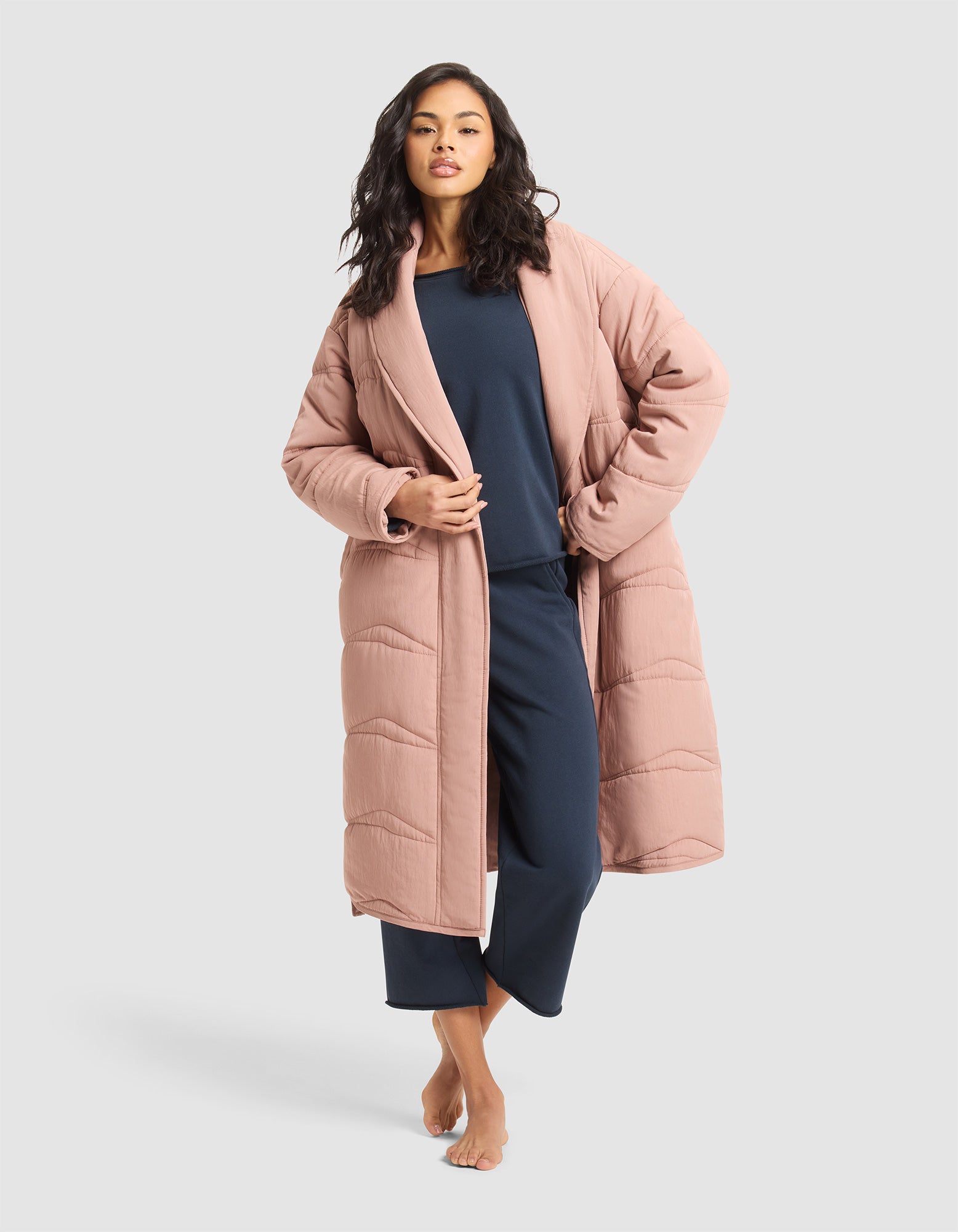 A woman with long wavy hair poses barefoot against a light gray background, wearing the Cozy Earth Women's Quilted House Coat in blush pink over navy blue loungewear. 
