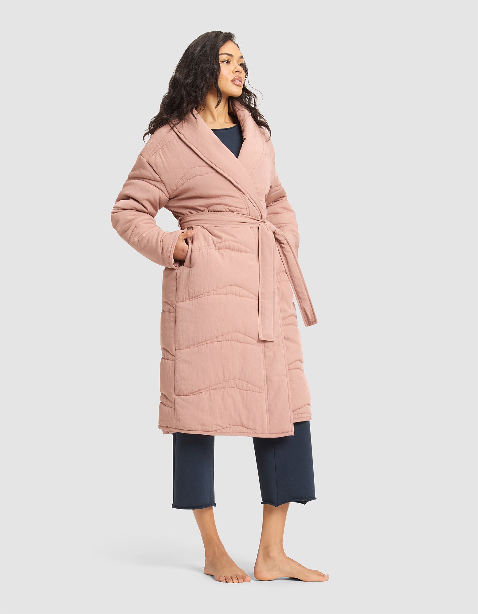 A woman stands barefoot in the Cozy Earth Women's Quilted House Coat, a long, light pink belted robe, layered over navy blue pajama pants and top, set against a plain light background. 