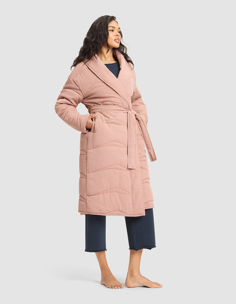 A woman stands barefoot in the Cozy Earth Women's Quilted House Coat, a long, light pink belted robe, layered over navy blue pajama pants and top, set against a plain light background.