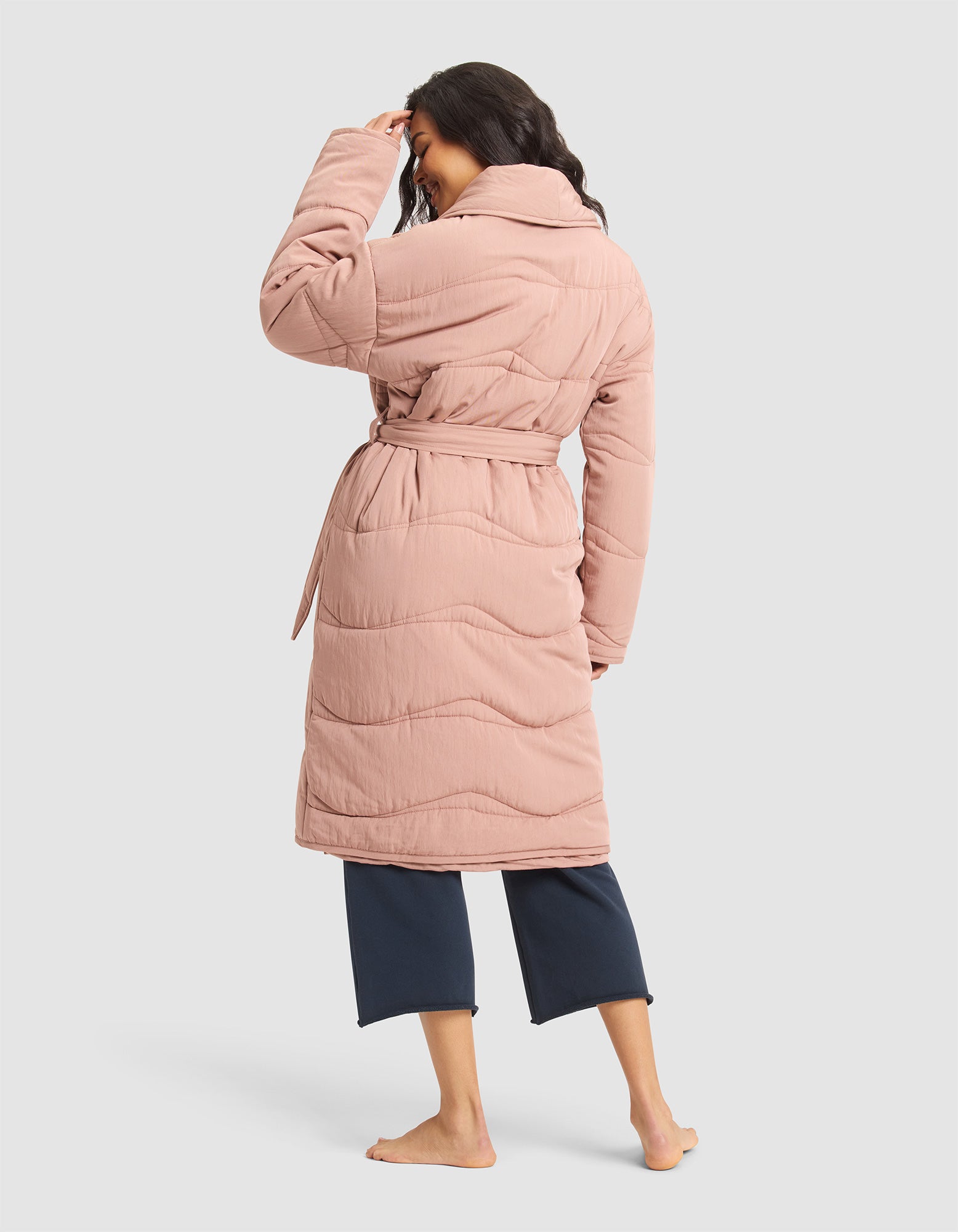 A woman stands barefoot, facing away, wearing the Cozy Earth Women's Quilted House Coat in light pink with a belt and wide collar over dark cropped pants, set against a plain light background. 