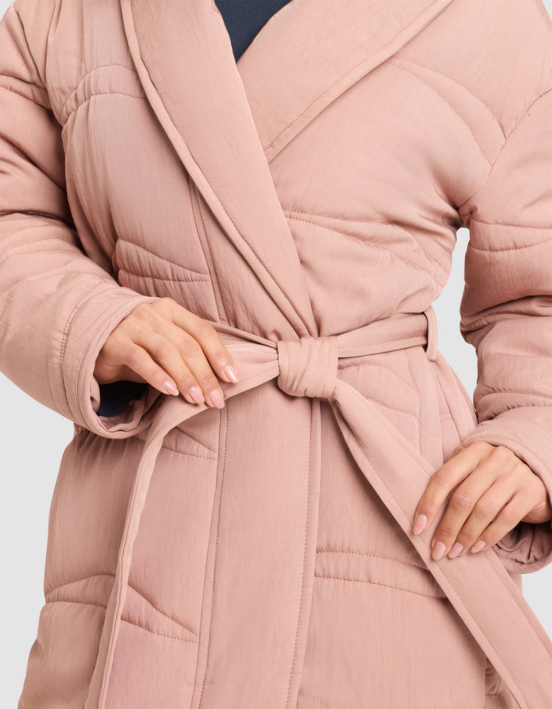 A person ties the waist belt of a Cozy Earth Women's Quilted House Coat in light pink, showing neatly manicured nails. The image highlights the hands and torso.