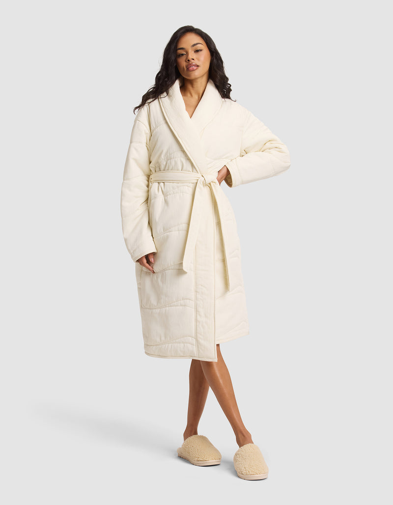 A woman wears the Cozy Earth Women's Quilted House Coat in Vanilla, featuring a waist tie and matching slippers, as she stands facing forward against a plain light gray background.