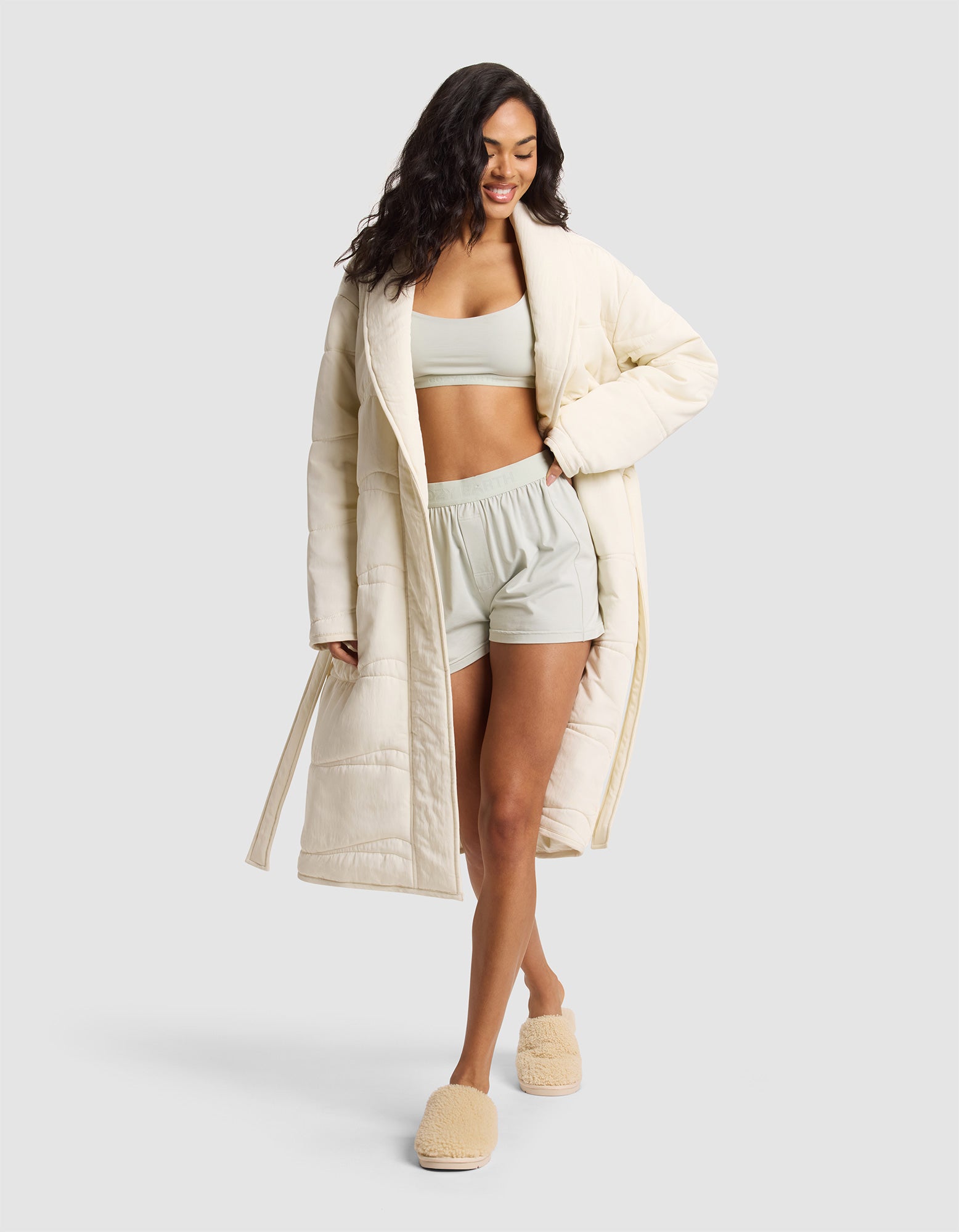 A woman smiles with one hand in her pocket, wearing the Cozy Earth Women's Quilted House Coat over a light sports bra and matching shorts, paired with fuzzy beige slippers, set against a plain light background. |Color:Vanilla