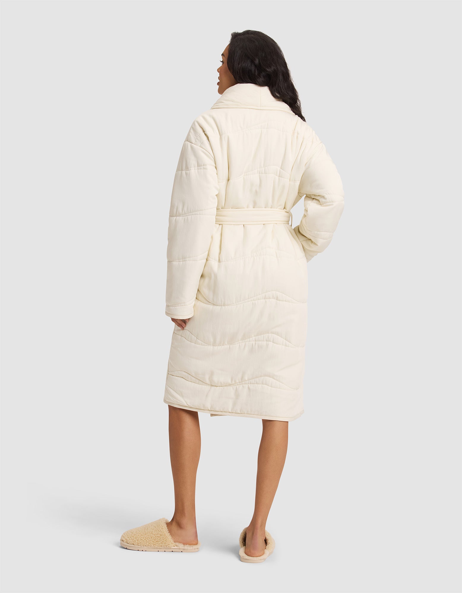 A woman faces away, wearing the Cozy Earth Women's Quilted House Coat in cream with a belt and tan slippers, set against a plain light gray background. |Color:Vanilla