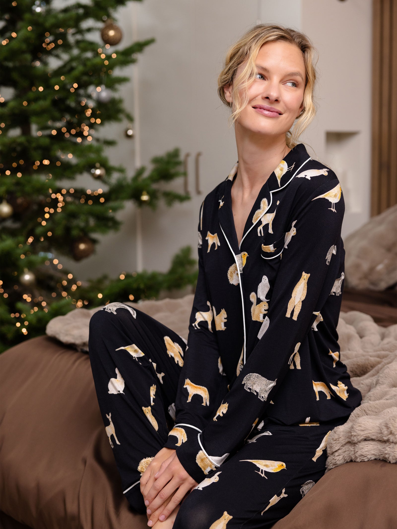 Woman sits by a Christmas tree wearing Women's Long Sleeve Bamboo Pajama Top in Stretch-Knit - Woodland Creatures Black 